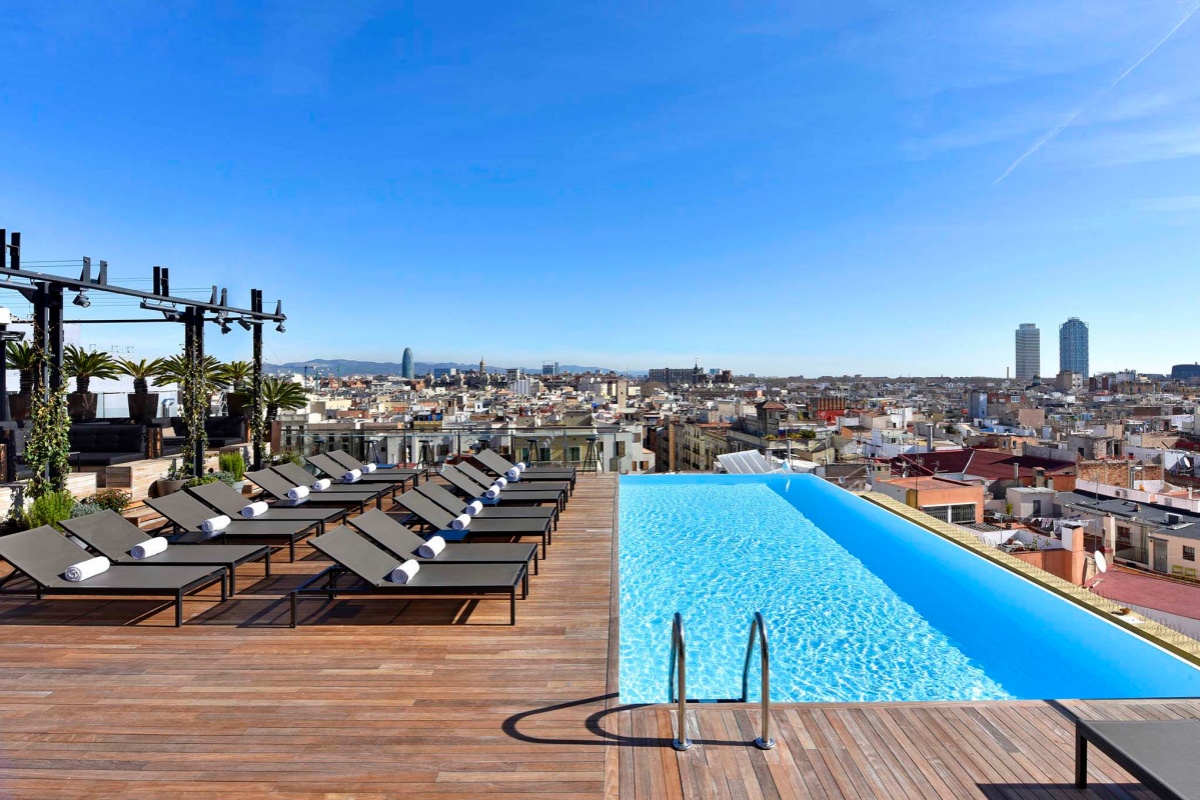 Grand Hotel Central - a pool on a rooftop with chairs and a city in the background